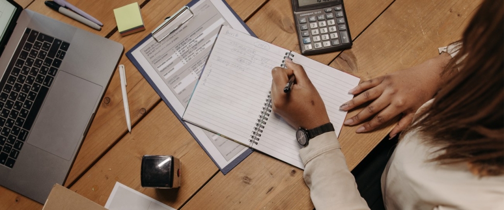 Top-down picture of woman sitting at a table, writing in a notebook, surrounded by documents, a laptop and calculator.